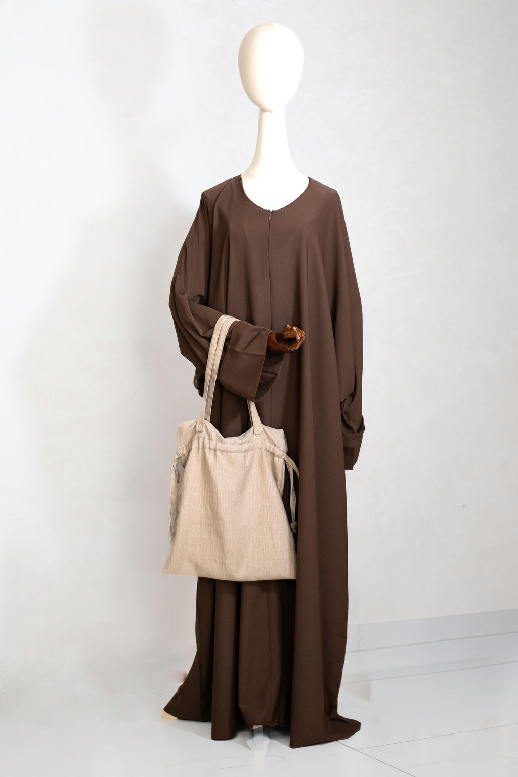Earth Brown abaya van Saahil Collection op houten mannequin met beige linnen tas, minimalistische achtergrond.